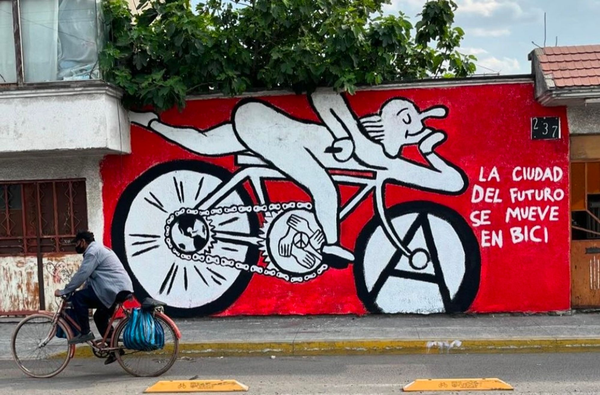 A graffiti of a crudely-illustrated person riding a bicycle spray-painted on a wall.

The front wheel of the bicycle has a large letter A (symbol of anarchism) inside. The bicycle chain on the back wheel wraps around an illustration of the globe. The front part of the chain in the middle contains an illustration made up of hands grabbing each other in a circle around the peace symbol.

The person riding the bike is all white with black outlines, and the wall behind it is deep red.

There is a person riding a bicycle in front of the wall with the graffiti. 