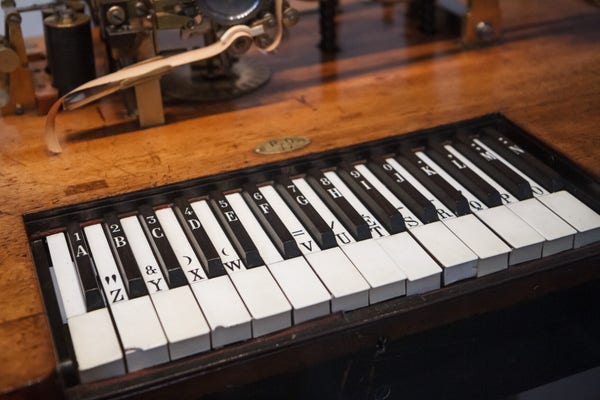 Old piano-style keyboard with keys marked with letters, numbers and punctuation symbols in a polished wooden desk, with bits of mechanical gadgetry partially visible above.