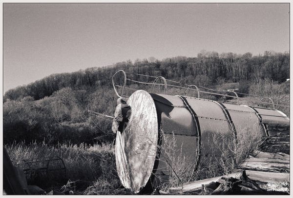 A big riveted and dented metal silo lies discarded on its side as if it were an empty beer can tossed into the bushes. The stair guard runs along the top side, there's a hatch on what used to be the top. In the foreground a loose stack of firewood, in the background rolling wooded hills.