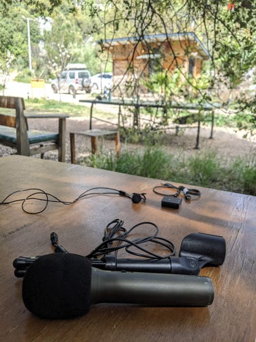 Full-size dynamic microphone with holder and cable in front of the table, small lavalier mic and tiny flash-drive sized audio interface at the other part of the table, beautiful mountains on the background.