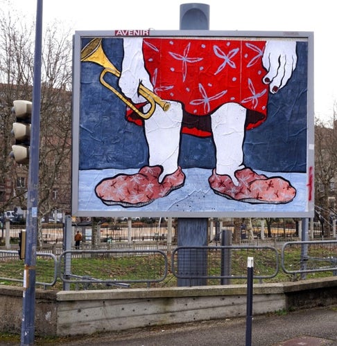 Streetartwall. Photo of a section of street with a billboard. On the billboard is a painted image showing the lower part of a woman in slippers; the painting style is crude and reminiscent of satirical depictions. The lady stands in pale pink slippers, has white legs, wears a red and blue patterned skirt and holds a golden trumpet in her oversized hand. The background is blue wallpaper. (In the photo there is a metal fence in front of the wall poster, a traffic light and houses behind it).
Info: The large billboard is part of a series of artworks by the artist duo Ella/Pitr in public spaces in St Etienne