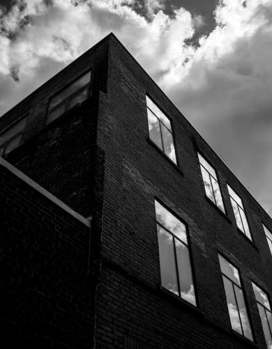 Looking up at a brick building covered in windows. The clouds that fill the sky can be seen reflecting in the shiny glass in this moody black and white image.
