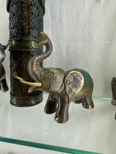 A brown Indian elephant made of metal with a carpet embossed on its back. The carpet is painted greed. Teeth, edge of ears, eyebrows and toe nails are painted in gold.