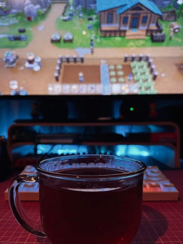 A clear glass mug of rooibos tea on a table with a blurred video game (Coral Island) on a screen in the background.