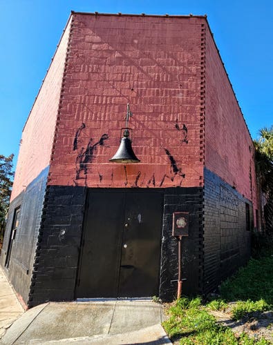A curiously shaped, brick building, long vacant, recently painted in thick, almost glossy black and maroon, covering the once vibrant artwork and graffiti.