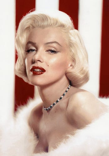 Her hair is blond, short and straight. She is wearing make up, including a red lipstick that highlights her lips. She is also wearing necklace.