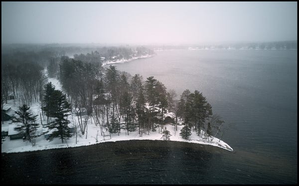 Aerial photo taken just about the tree line along the shores of an open lake, during a snow storm. The ground and cabins are all covered in snow. The sky is dark and snow can be seen in the air.