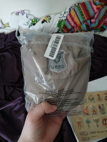 My hand holding up a brown hat with a silver patch that reads "UESP" that's still inside its plastic packaging.