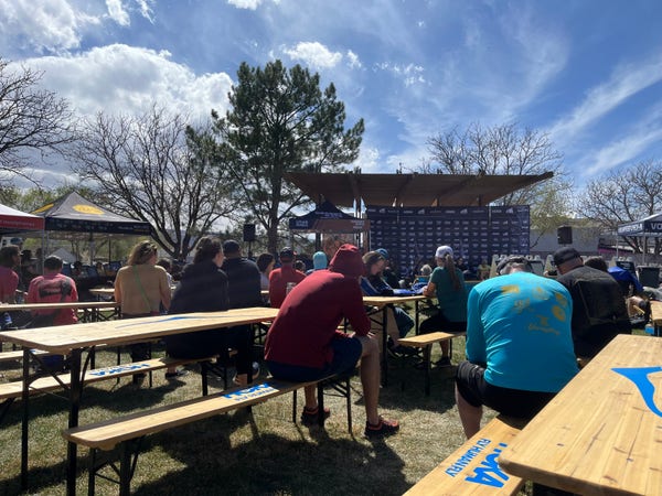 Desert Rats 100k brief, staff in the background runners sitting in wooden benches all in the outdoors.