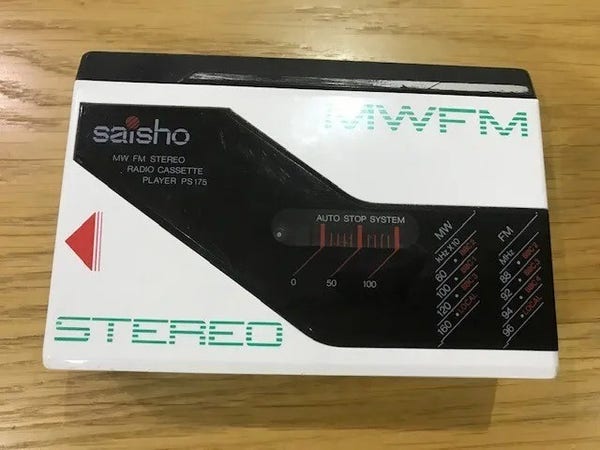 A Saisho Walkman-type personal radio cassette player. It's white with green lettering in pseudo-computer or TV scan lines style reading MWFM top right and Stereo bottom left. A black flash from top left to bottom right includes the brand name and the MW/FM Stereo Radio Cassette plater letting, some MW and FM bands, and a space to see the tape inside.