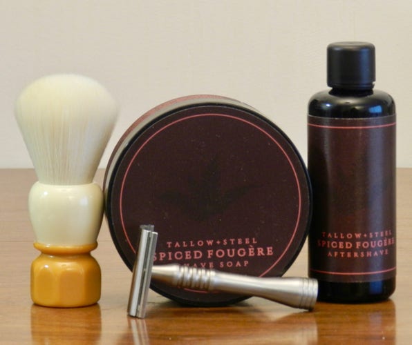 A shaving brush with white synthetic knot, white hand, and butterscotch octagonal base stand next to a pub of shaving soap with a black label with a narrow red ring near the edge. In red letters at the bottom is printed "Tallow + Steel / Spiced Fougère / Shaving soap." Next to it is a narrow cylindrical black bottle with a black cap and the same label except that the bottom line on this one reads "Aftershave." In front is a matte-finish stainless steel DE razor whose handle is ribbed at the top and bulges at the base.