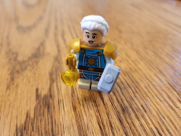 The female dwarf cleric from the Lego Dungeons & Dragons set. She's a small-sized minifigure with shorter legs, with blue and tan painted garb on the body pieces. She has a separate piece representing bronze-colored pauldrons. She has a warhammer in her left hand, and a piece representing her holy symbol in her right. This version of the figure has an amused expression.