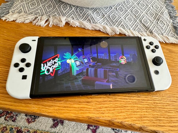 Photo of a Nintendo switch with white controllers, the screen shows a gaming room with a juke box, arcade machines, and a “we’re open” sign with someone sat playing a machine.   