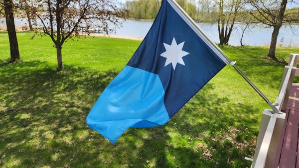 A new Minnesota flag featuring 8pt star flies at a home against green grass and a blue lake.