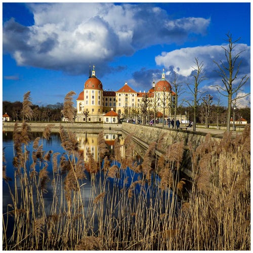 Baroque castle in Moritzburg/ Saxony reflected in a lake, surrounded by trees and dried reeds under a cloudy blue sky.