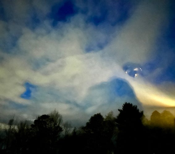 Cloudy night sky with glimpses of blue and an odd yellow shaft with a familiar “ufo” hovering in the sky from reflection of ceiling lamp. 
