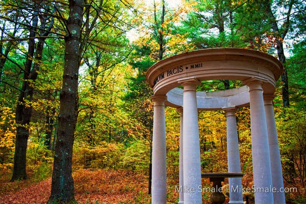 A Greco-Roman styled circular temple with doric pillars sits beneath an enclave of pine trees. Deciduous trees, green with gold colours coming in sit around the structure in the background. A pile of red, orange, and brown leaves lie on the forest floor in the bottom left and behind the temple.