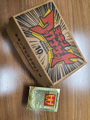 Photograph of a box of 10 chicken McNuggets from McDonald's with a sauce packet next to it with a gold lid that says "WcDonald's Sauce." The McNugget box is a light brown box with bold Japanese lettering in red that reads "Chicken WcNuggets" - the English language translation is underneath. The front side of the box reads "10 Chicken McNuggets" in both Japanese and English.