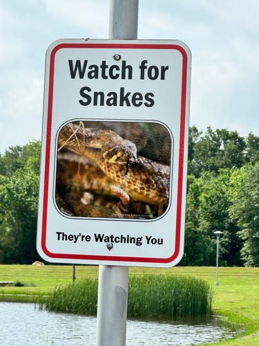 A warning sign reading "Watch for Snakes - They're Watching You" with an image of a snake, mounted on a post with a pond and trees in the background.