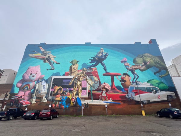 Cincinnati Toy Heritage Mural with characters from the old Kenner Toy company. Some of the characters are: C-3PO from Star Wars, various toys from the movie Toy Story like Mr. Potatohead, Care Bears, the Ghostbusters hearse, Easy Bake Oven, Spirograph and more painted on a large brick building adjacent to a paid parking lot. 