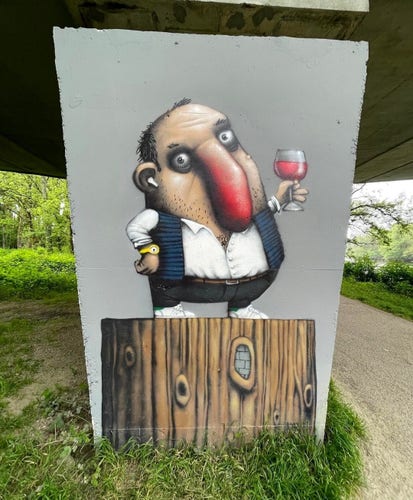 Streetartwall. A mural of a hard-drinking cartoon character has been spray-painted on the gray concrete wall under a bridge. A strange little man in black pants, a white shirt and a sleeveless black jacket is standing on a tree stump with a glass of red wine and toasting us. He's half bald, has a huge red nose (which looks a bit like a genitalia) and dark circles under his eyes. He wears a headset in his ear, a gold watch on his arm and his chest hair goes straight into a three-day beard. The photo shows grass and a path with a riverbank on the right.