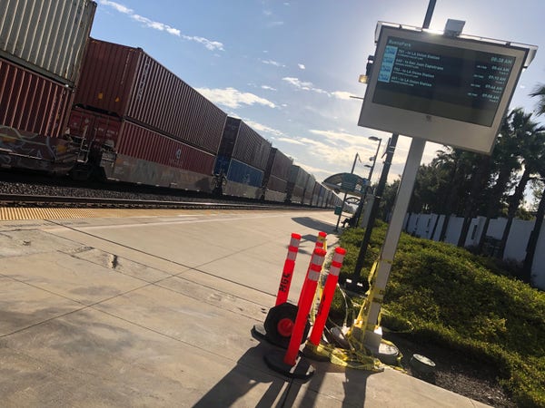 I’m standing on an tempts platform waiting for the train to arrive. The center track has stationary freight cargo with double stacked containers. The sky is blue with some white clouds. The bright sun is hidden behind a train o for display with the next few trains listed.