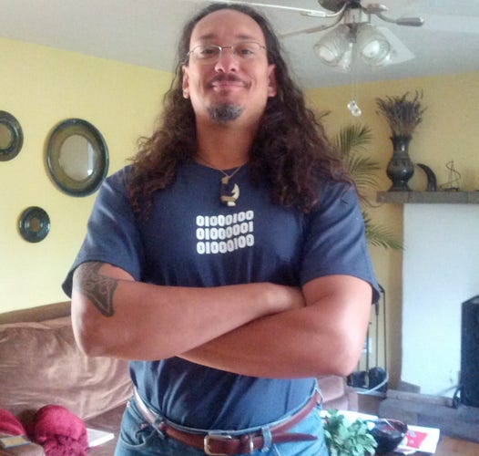 A large, well-muscled light-skinned Black man with long hair, a mustache and goatee. He's wearing a blue shirt with "DAD" written in binary code and has a Maui hook around his neck. His arms are folded showing fairly sizable arms.