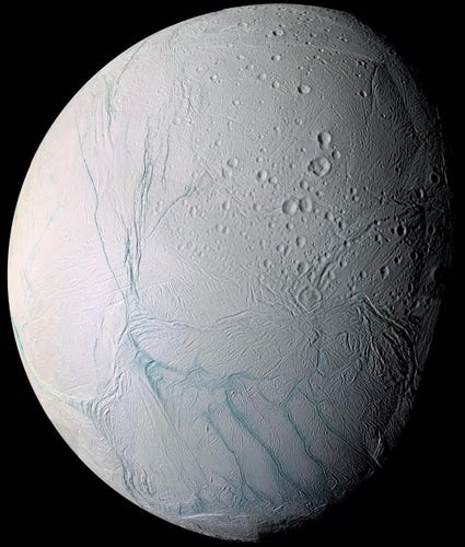Close-cropped photo of Enceladus, silhouetted brightly in dark space. It’s mostly white with sporadic streaks of light blue. The surface is wrinkled with a number of deep craters.