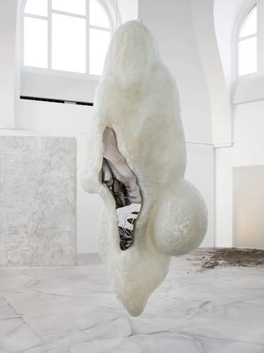 Hanging sculpture of a long, white, bulbous form with a curving opening slit with various material inside