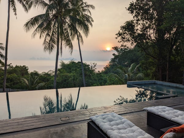 Sunrise over the ocean reflected in the ocean and the surface of an infinity pool. Palm trees are in the foreground and lush greenery is between the pool and the ocean below. Two lounge chairs await beside the pool.