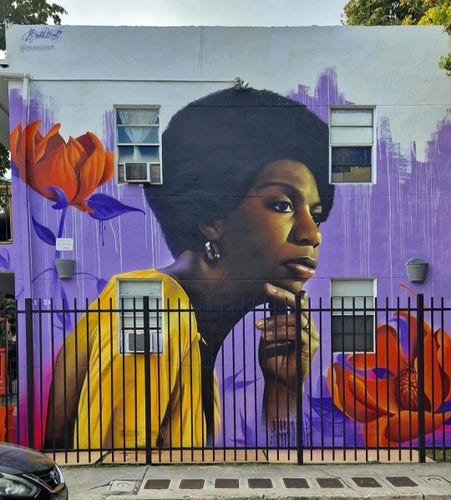 Streetartwall. A beautiful portrait of the black singer Nina Simone was sprayed/painted between four windows on the outside wall of a single-storey modern house. She is depicted up to her chest and looking thoughtfully to the right. One hand is under her chin. She has short black hair and is wearing a yellow dress. The background is purple with two large red flowers on it.  A beautiful and realistically designed tribute.
Info: Nina Simone (real name Eunice Kathleen Waymon; * February 21, 1933 / † April 21, 2003) was an American jazz and blues singer, pianist, songwriter and civil rights activist.