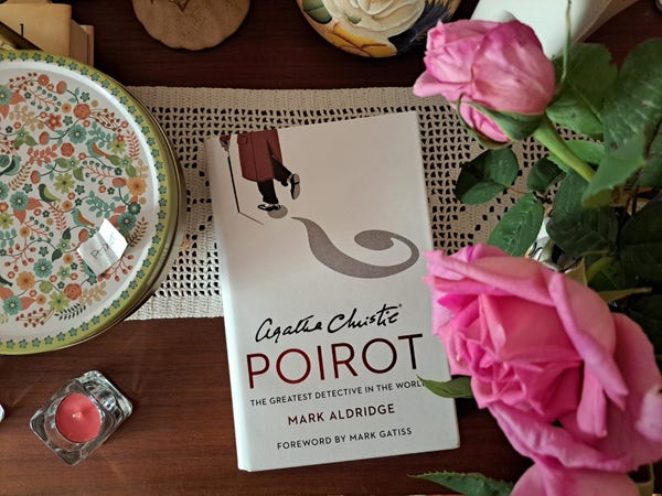 The book Poirot The Greatest Detective in the World by Mark Aldridge with a white cover with red title and a drawing of Poirot with a shadow in the form of an question mark on a wooden table next to a bar with pink roses a red candle and a tin with nature drawings in green, blue, and orange with a little scrap of paper that says Poirot