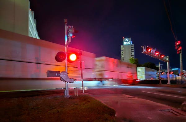 From the sidewalk in front of the flashing red crossing guard arms of a railroad crossing a freight train speeds past in the dark night.