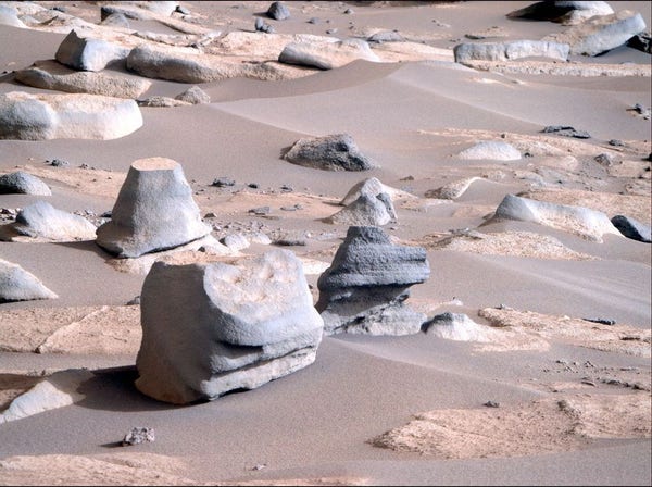 Numerous interesting rocks, scarred by time and wind (and maybe water) are pictured on the surface of Mars, resting on the lighter coloured sand.