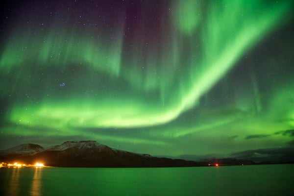 A night scene. Stars dot the night sky not hidden by a huge aurora display. Green swirling light dominates the sky. Reflecting in the ocean and painting the mountains in the distance. 
