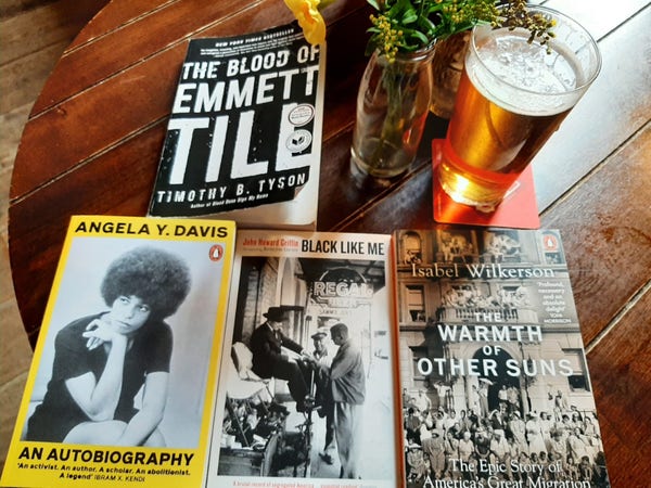 Clockwise from top left, The Blood of Emmett Till by Timothy B. Tyson; A yellow flower in a smoothie bottle; a pint of Fuller's Grand Slam Spring Ale; The Warmth of Oyher Suns by Isabel Wilkerson; Black Like Me by John Howard Griffin; An Autobiography by Angela Y. Davis.