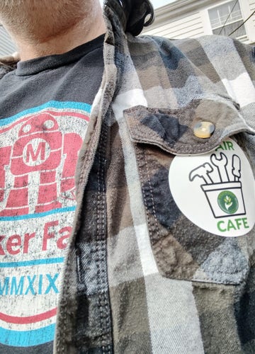 Closeup of the chin and chest of an unkempt middle-aged guy wearing a Maker Faire T-shirt, a grey plaid flannel, and a black and green circular Repair Cafe sticker.