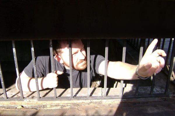 Photograph of a light skinned, red bearded man in a grey t-shirt inside of a concrete / stone structure with jail bars. He is holding onto the bars with his right hand while squinting and reaching up / out with his left hand.