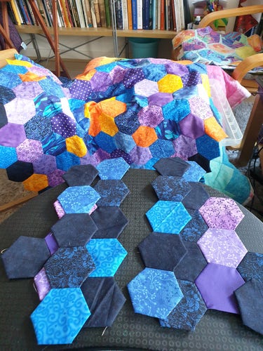 Photograph of uneven clusters of three inch fabric hexagons on a lap desk. There's are under preparation, and will be added to the big hexagon made of more hexagons which is draped in the background. The hexies are in various shades of blue and purple with some in bright yellow and orange.

In the background is a chair with a seat cover made of bigger hexagons appliquéd to some sky blue linen I salvaged out of a museum case lining many years ago. This makes it look like I love colour and I like hexagons, both of which are correct.  