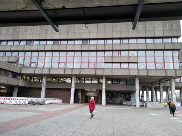Photo of the library on the Ruhr University Bochum campus. A (neon? Not glowing at the moment) sign just below the roof line, mirrored for some reason, reads "UNIVERSITATBIBLIOTHEKBOCHUM". The last letter or two are cropped out of frame and not visible.