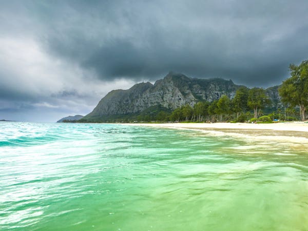 Turquoise water meets a sandy beach with trees; stormy clouds loom over a rugged mountain in the background.