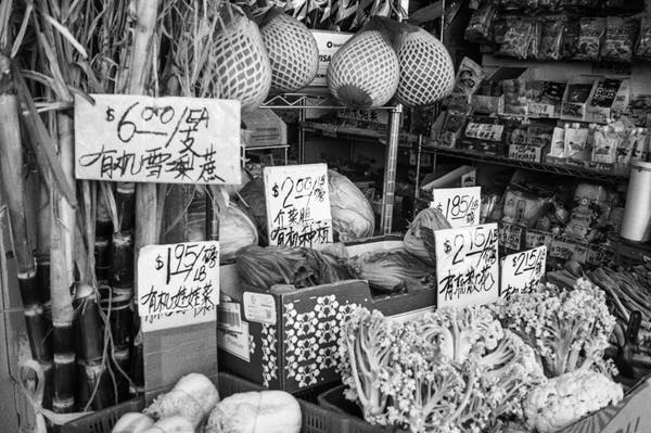 a scan of a black and white 35mm negative of a bunch of stuff like vegetables outside a chinatown shop, looking messy. there's a bunch of prices and labels in chinese
