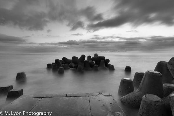 Black and white photo of a seascape with tetrapod breakwaters and dramatic cloudy skies.