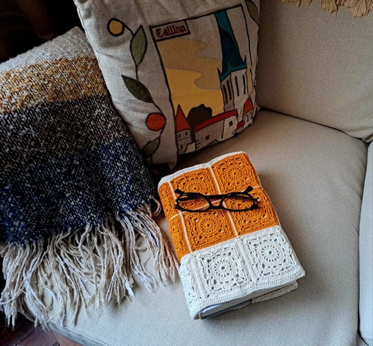 A blanket, a cushion, and book with glasses on an armchair. The book has a cover in crochet.