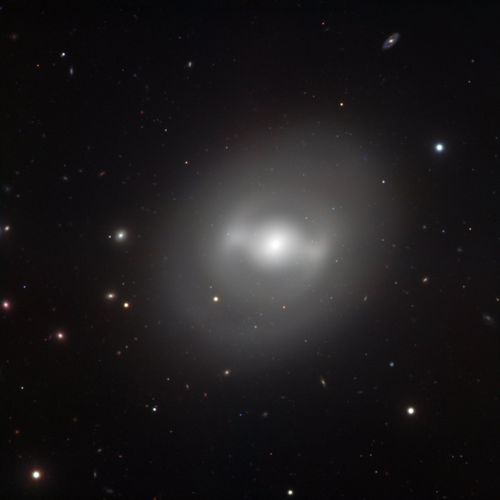 A telescopic image of a galaxy against a black background. The galaxy is a white, fuzzy elliptical cloud with a bright central core. There are two T-shaped bright structures emerging from opposite sides of the core.