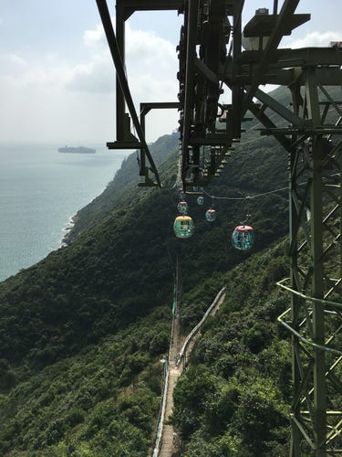 Sky gondolas going across a mountain with water to the left there is also a walking path 