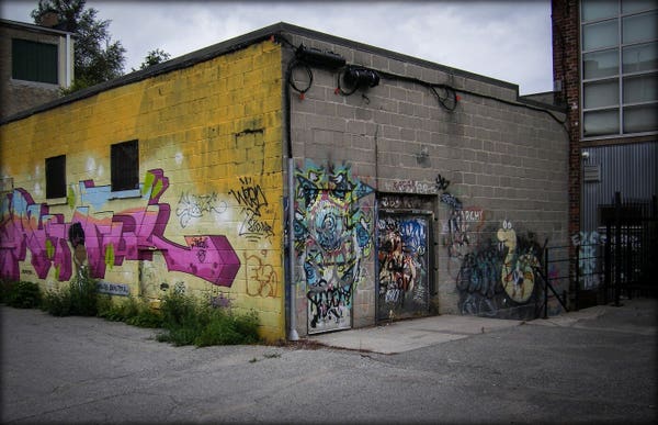 Graffiti covers two sides of a small building. One wall is painted a bright yellow, with a pink mural. The other side is covered in multi coloured artwork against the grey brick.