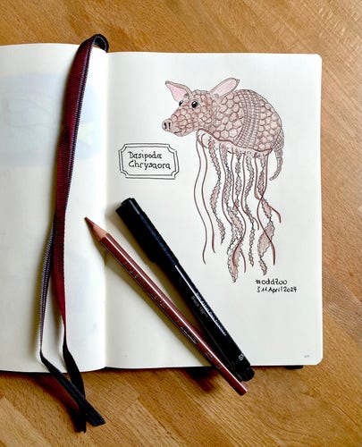 An open sketchbook with a drawing of a fictional creature that has the body of an armadillo and the tentacles of a jellyfish, next to colored pencils and a pen on a wooden surface. A label in the drawing reads "Dasipoda Chysaora“ and #OddZoo S 11.April 2024
