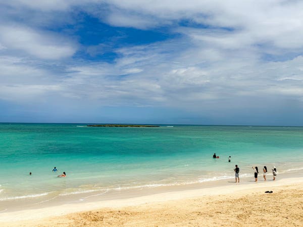 A sandy beach with people wading and swimming in turquoise waters under a blue sky with scattered clouds.