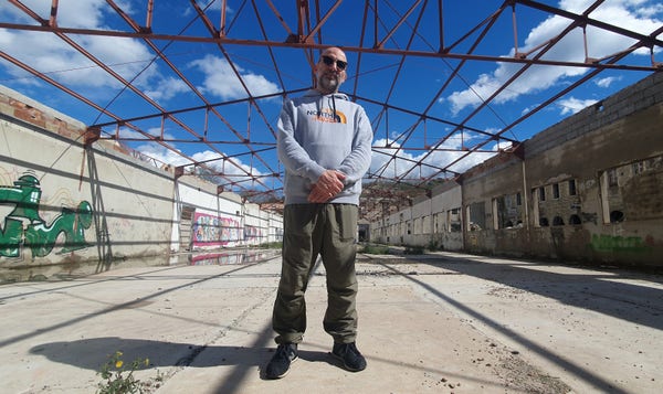 a middle aged man in sportswear poses in industrial ruins like he's about to drop a hot mixtape.